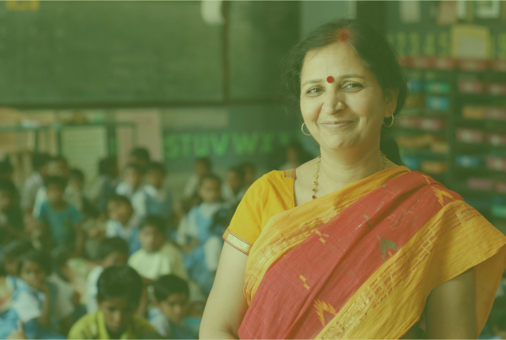 A woman wearing a brightly colored sari stands in a classroom, with blurred students seated in the background.