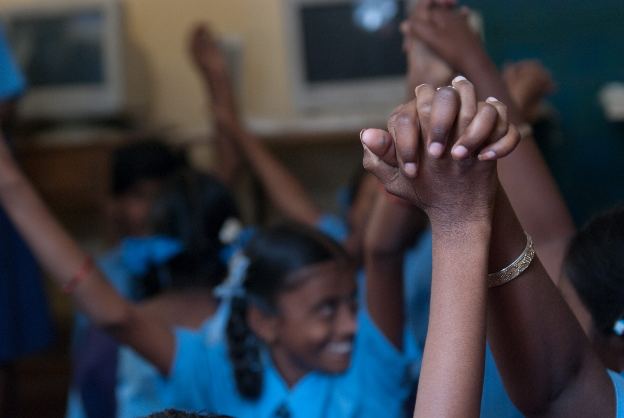 Students with hands raised, in the background. In the foreground are two hands raised, clasped together.