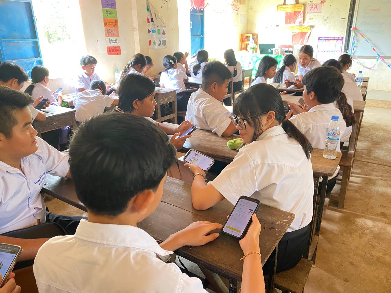 A classroom of students in white school uniforms sit at wooden desks, each using a smartphone during a lesson. The students are arranged in small groups and appear engaged with activities on their screens in a bright, sparsely furnished classroom with educational posters on the walls.